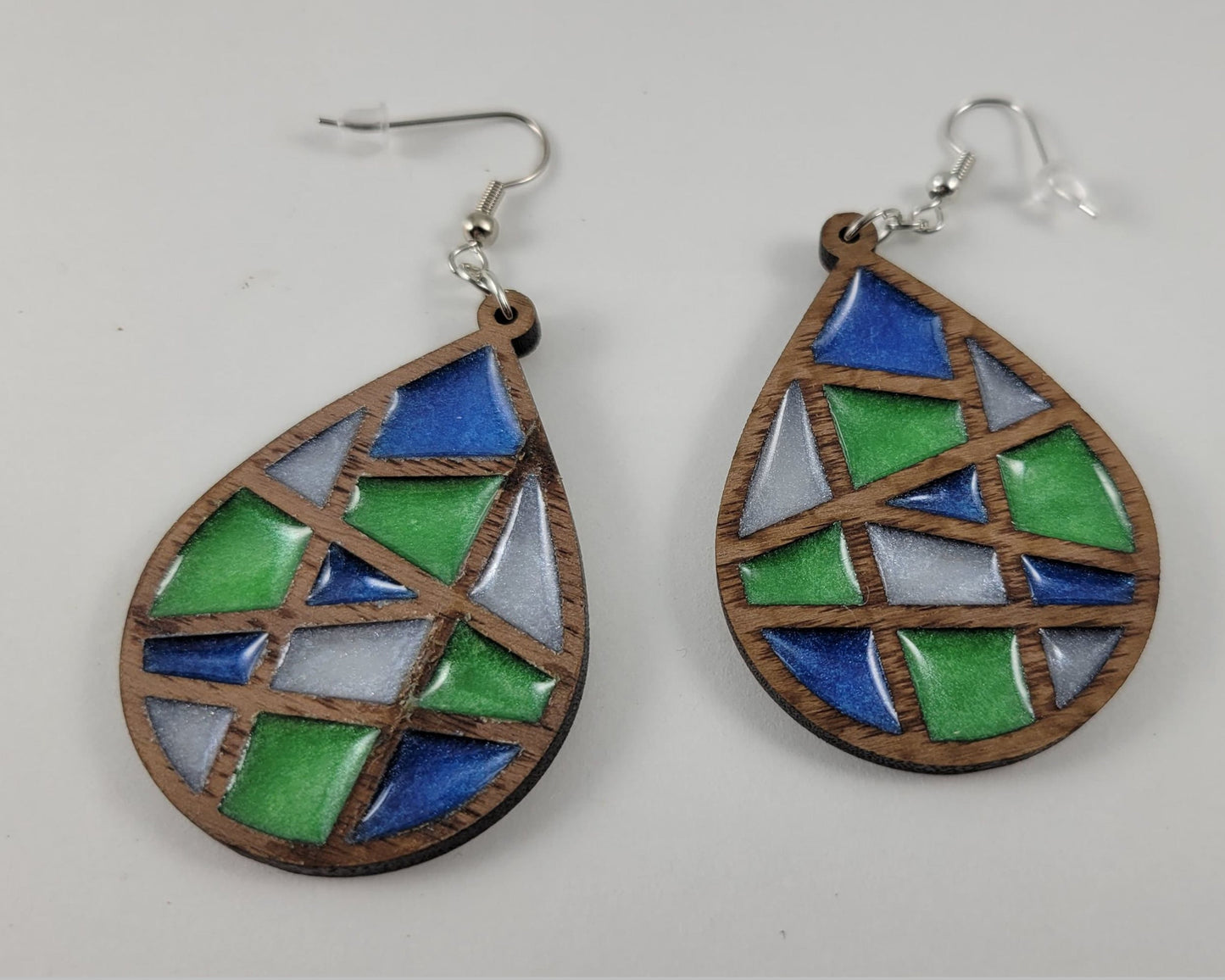 A pair of abstract-patterned earrings with a geometric design, featuring blue, green, and clear resin sections, with a wooden texture and silver hooks.