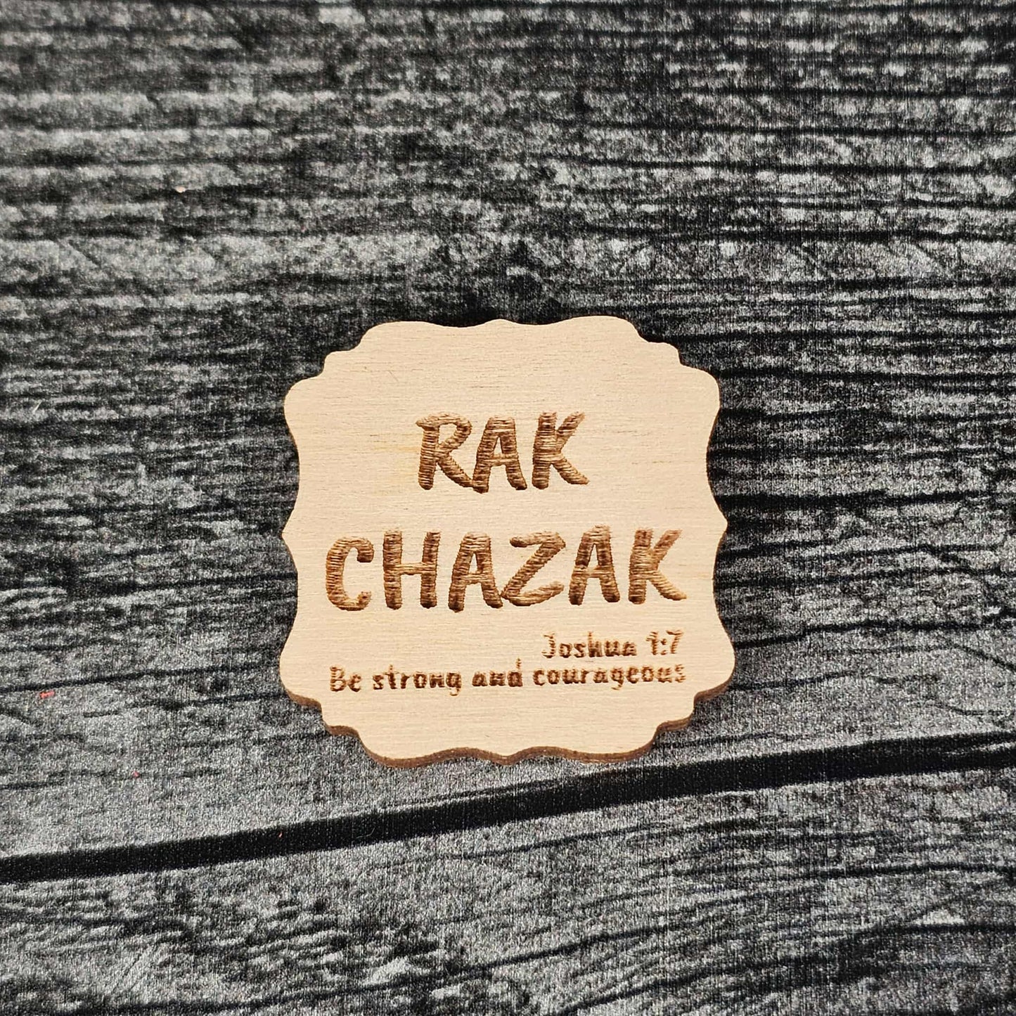 Decorative unstained wooden magnet with 'RAK CHAZAK' text, as well as a quote from Bible verse Joshua 1:7 on a textured surface.