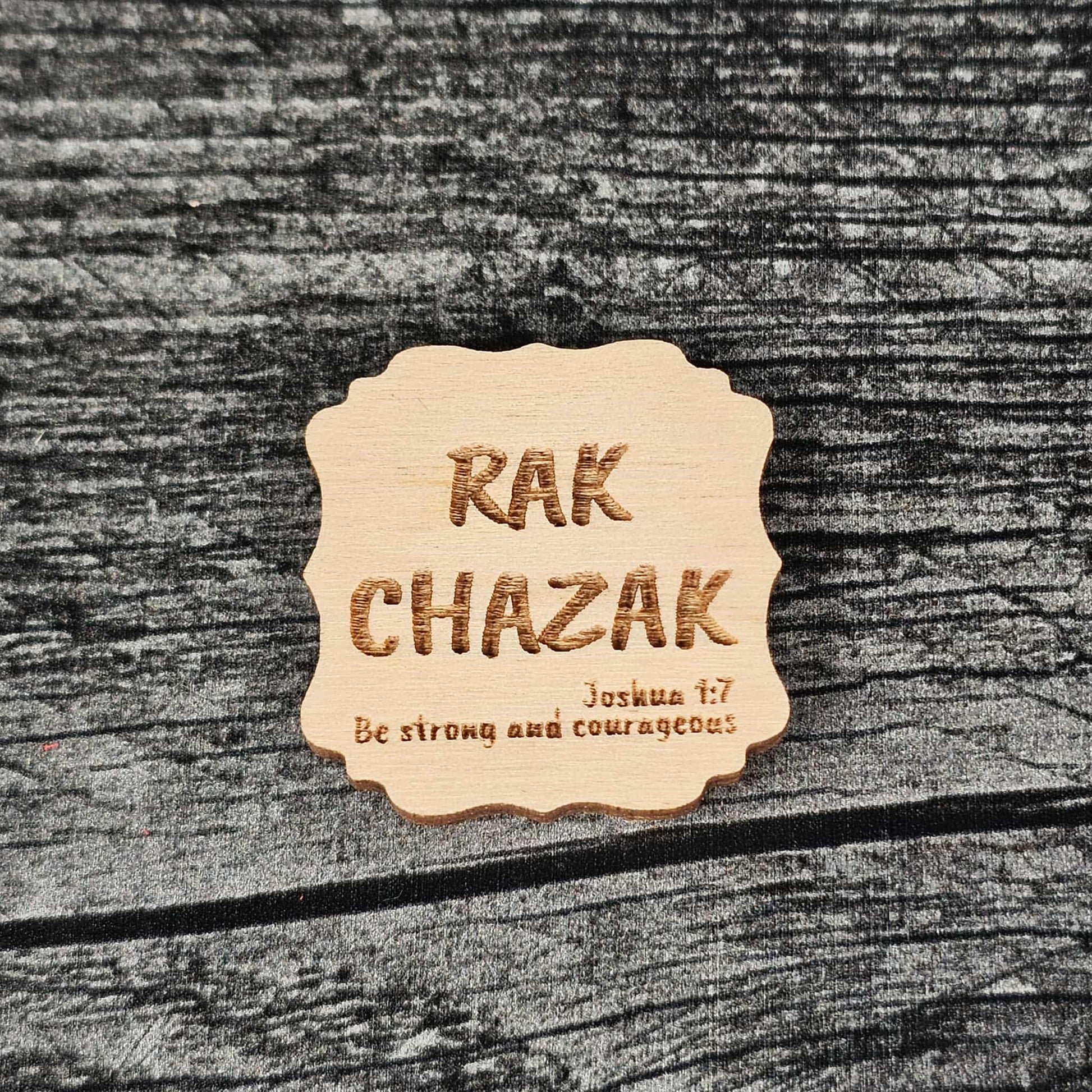 Decorative unstained wooden magnet with 'RAK CHAZAK' text, as well as a quote from Bible verse Joshua 1:7 on a textured surface.