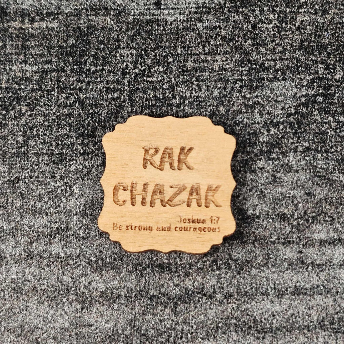 Decorative walnut stained wooden magnet with 'RAK CHAZAK' text, as well as a quote from Bible verse Joshua 1:7 on a textured surface.