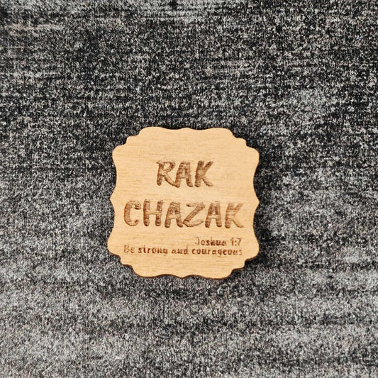 Decorative walnut stained wooden magnet with 'RAK CHAZAK' text, as well as a quote from Bible verse Joshua 1:7 on a textured surface.