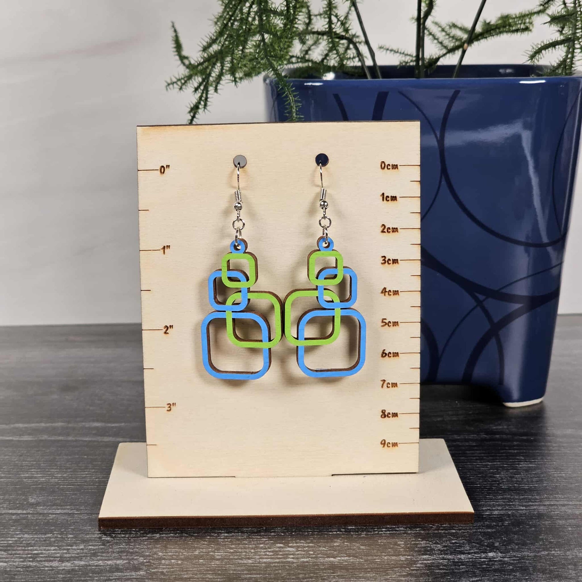 Geometric blue and green earrings on a wooden display card with measurement scale, against a neutral background.