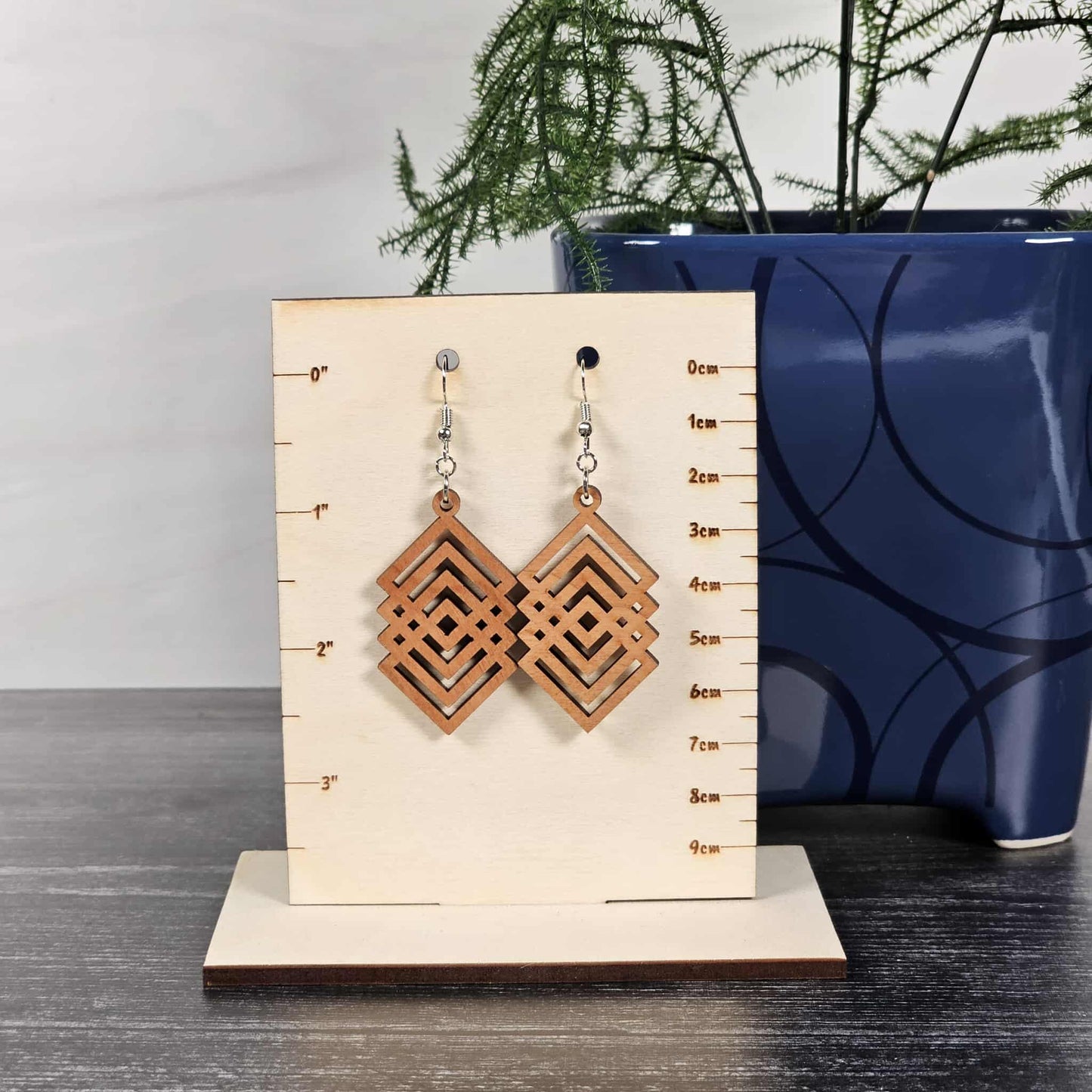 Pair of cherry geometric diamond earrings on a wooden stand with measurements, against a neutral background.