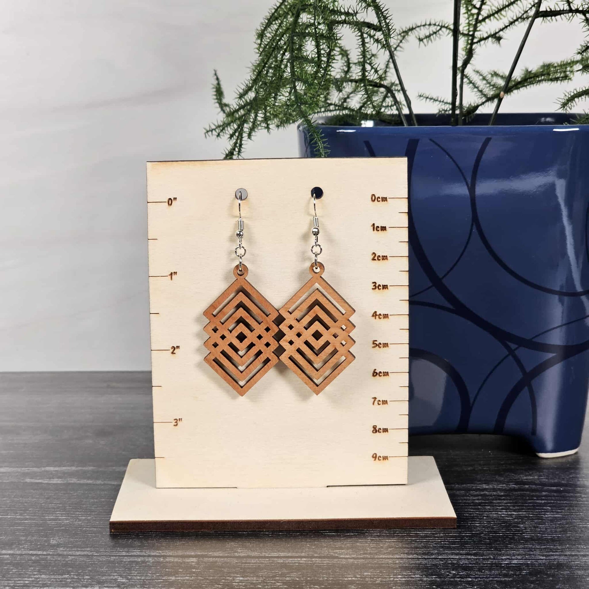 Pair of cherry geometric diamond earrings on a wooden stand with measurements, against a neutral background.