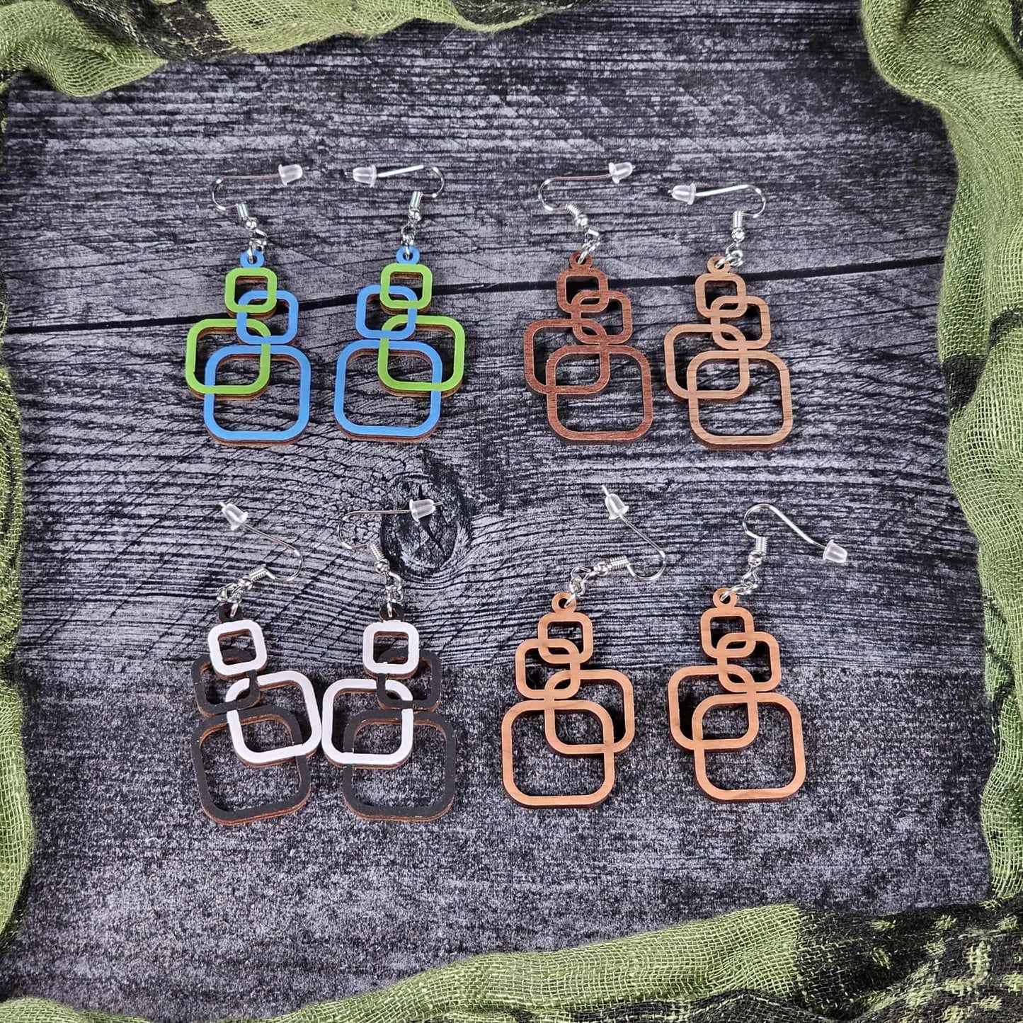 Group shot of the geometric squares earrings on a wooden surface with green fabric around.  All the color options are shown (black and white, blue and green, cherry, and walnut)