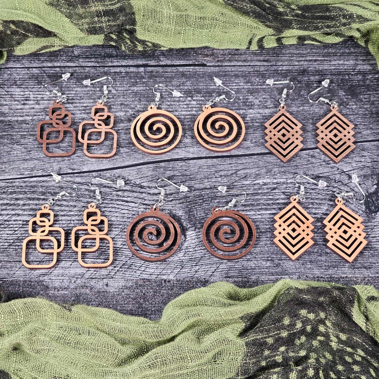 Group shot of all the different designs of the earrings (walnut and cherry options) on a wooden surface with green fabric in the background