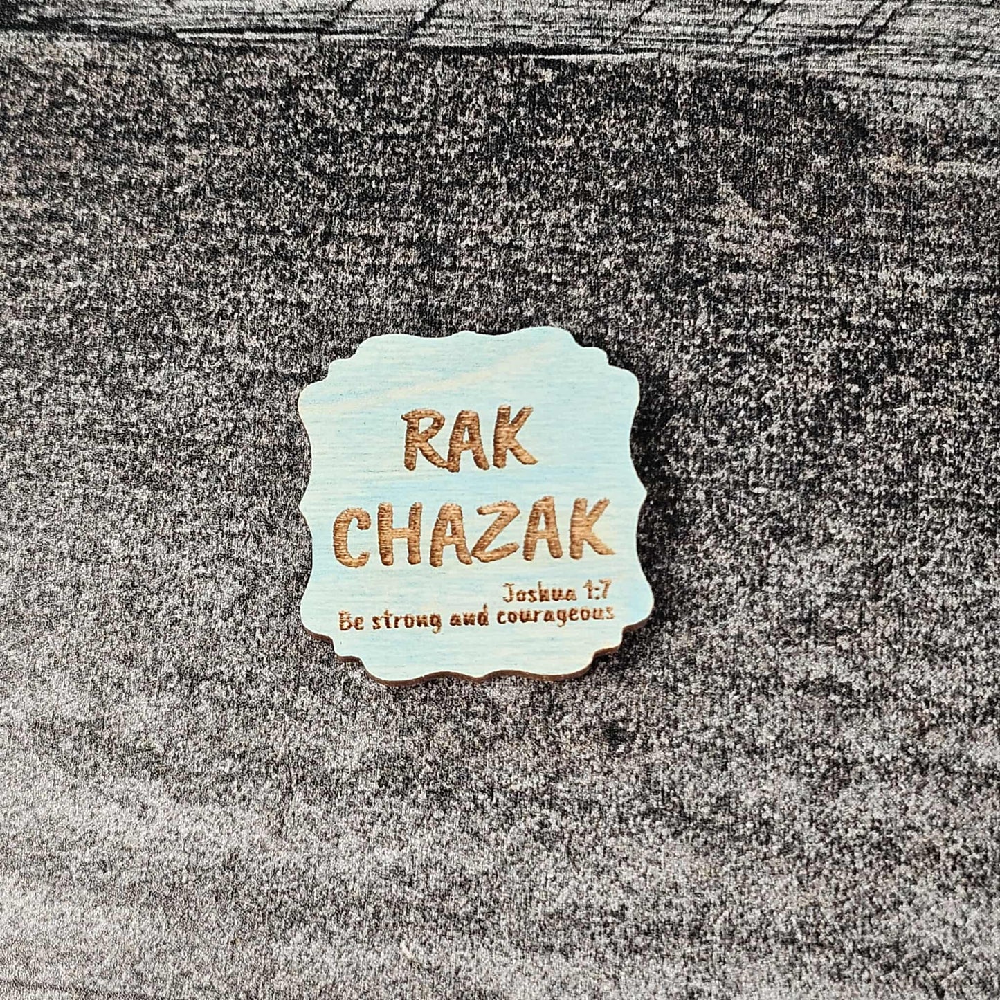 Decorative blue stained wooden magnet with 'RAK CHAZAK' text, as well as a quote from Bible verse Joshua 1:7 on a textured surface.