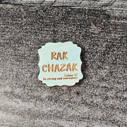 Decorative blue stained wooden magnet with 'RAK CHAZAK' text, as well as a quote from Bible verse Joshua 1:7 on a textured surface.