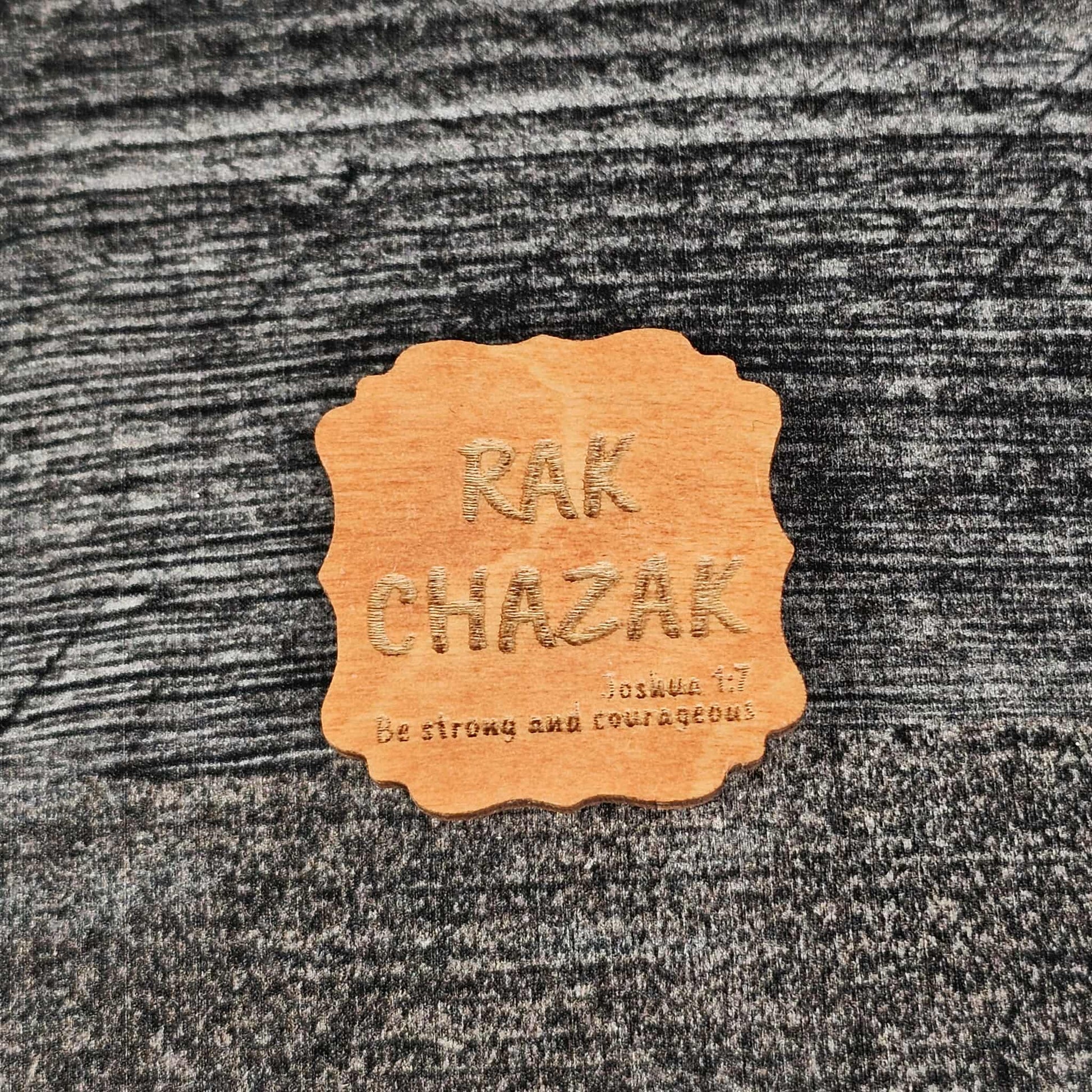 Decorative gunstock stained wooden magnet with 'RAK CHAZAK' text, as well as a quote from Bible verse Joshua 1:7 on a textured surface.