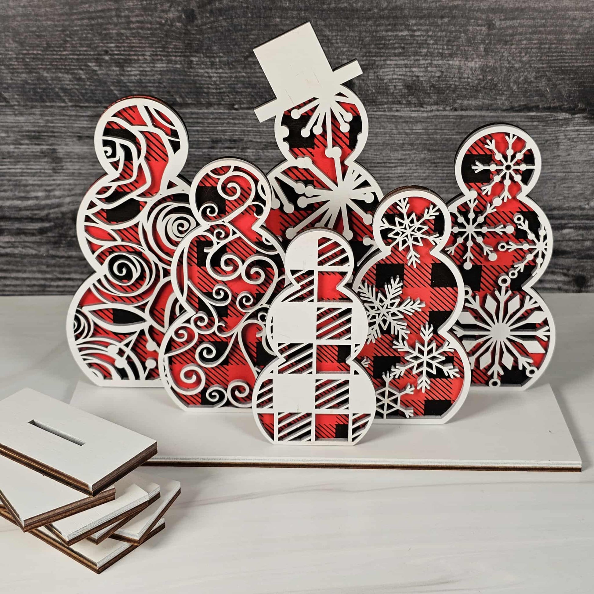 Decorative wooden cutout with red and black buffalo plaid color and white snowflake patterns on a wooden surface.