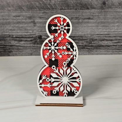 Wooden snowman decoration with red and black buffalo plaid pattern topped with a snowflake design in white standing on a wooden background