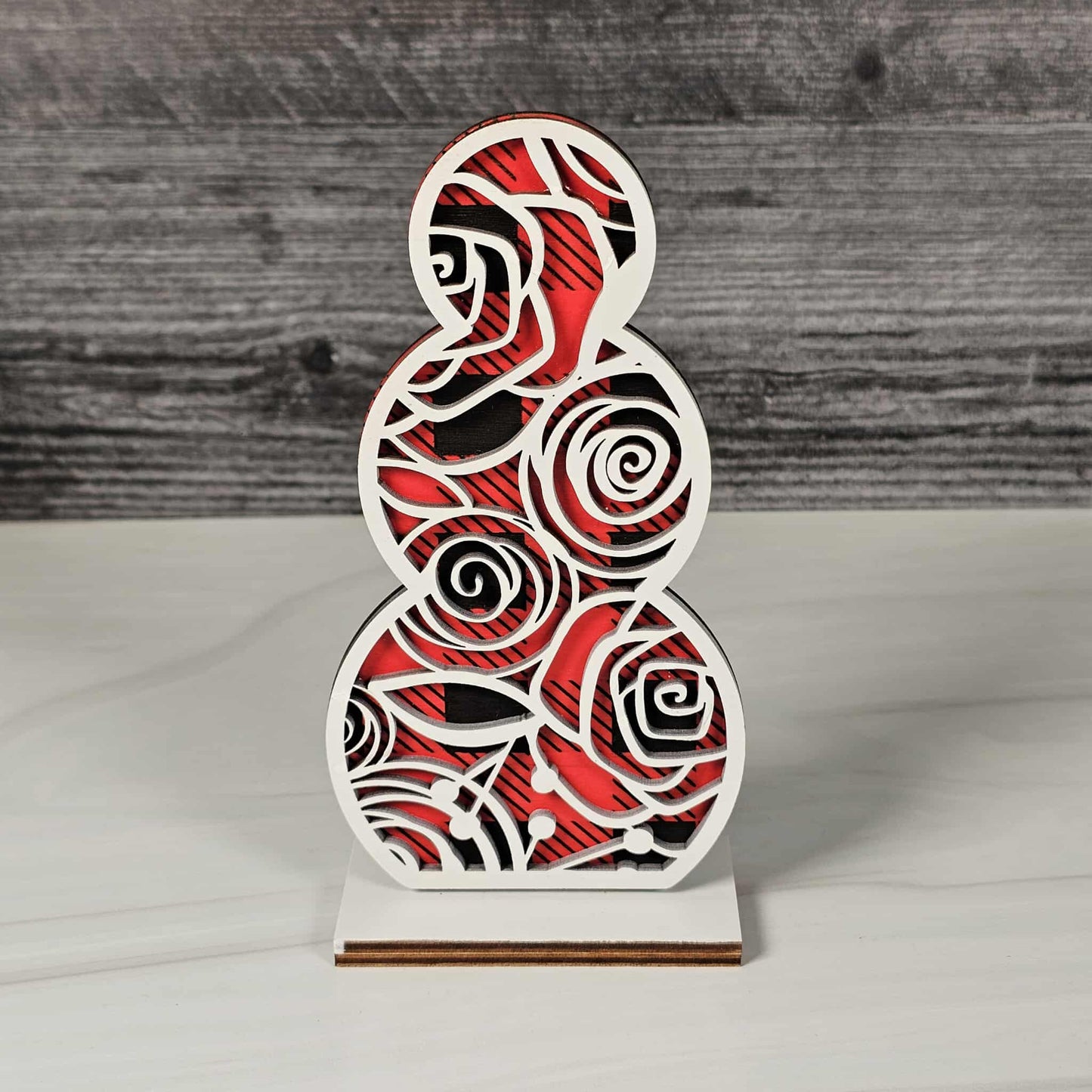 Decorative wooden snowman sculpture with red and black buffalo plaid pattern topped with swirly rose design in white 