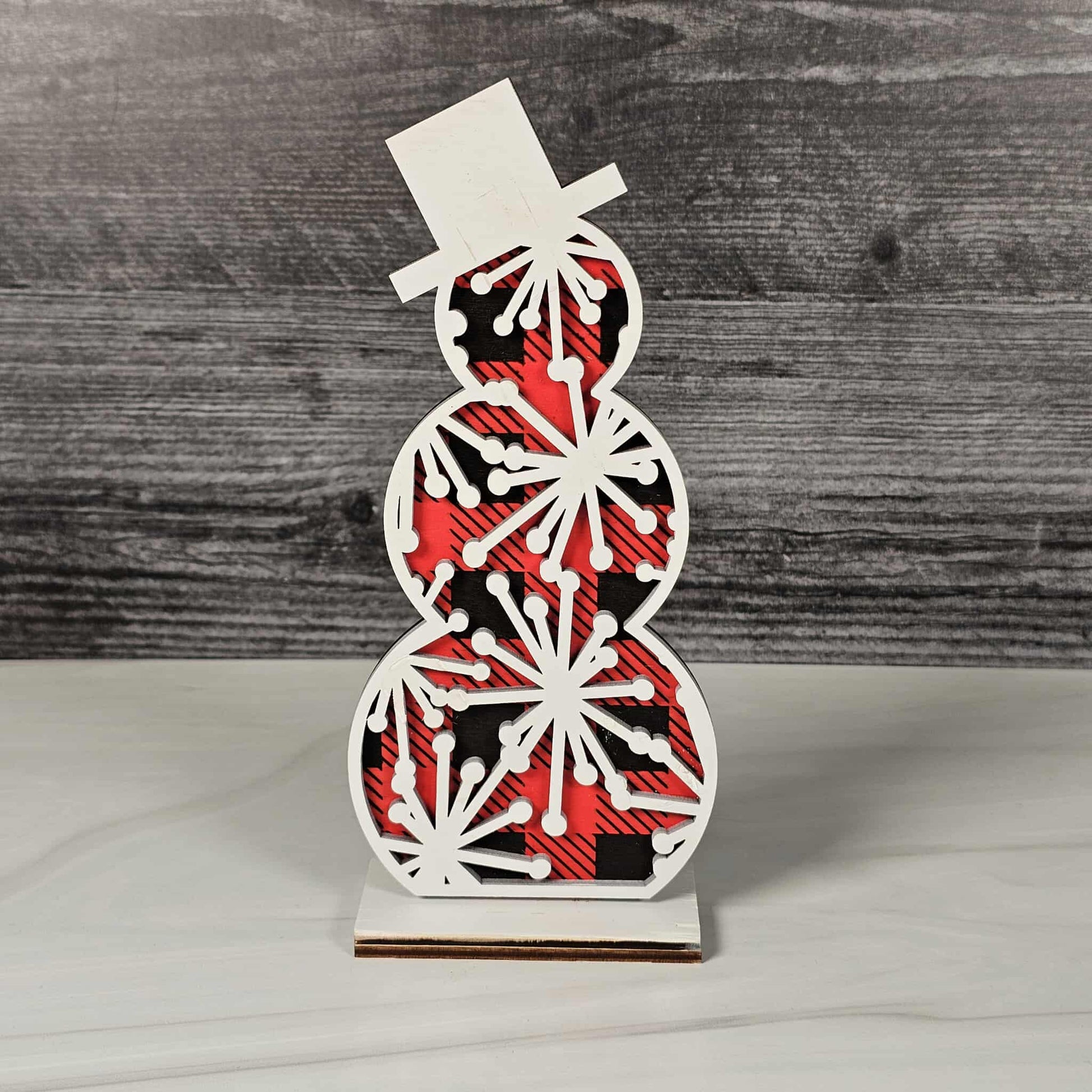 Decorative snowman figure with red and black plaid pattern on a wooden background with white starburst snowflakes on top