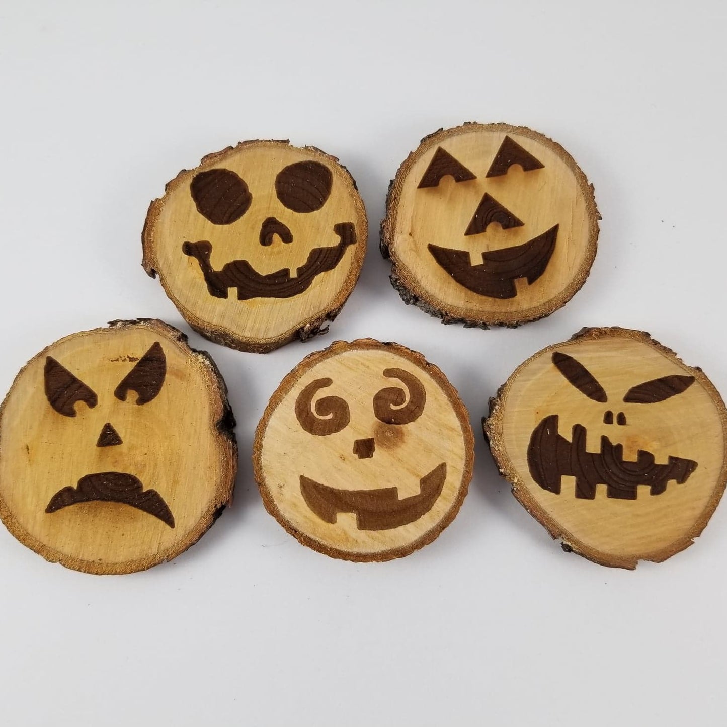 A set of five pumpkin face magnets of varying sizes, with different expressions such as happy, sad, and scared, laser engraved on a wooden surface.