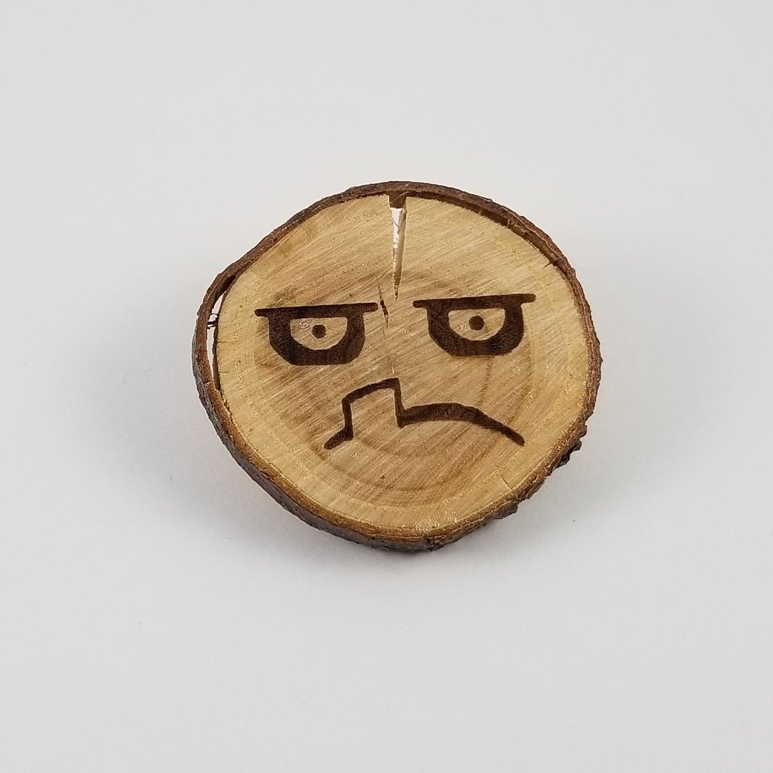A wooden toy figure with a cartoonish face engraved on it, resembling a gremlin or a gnome, with a sad expression. It has two faces, one on each side, for different viewing angles.