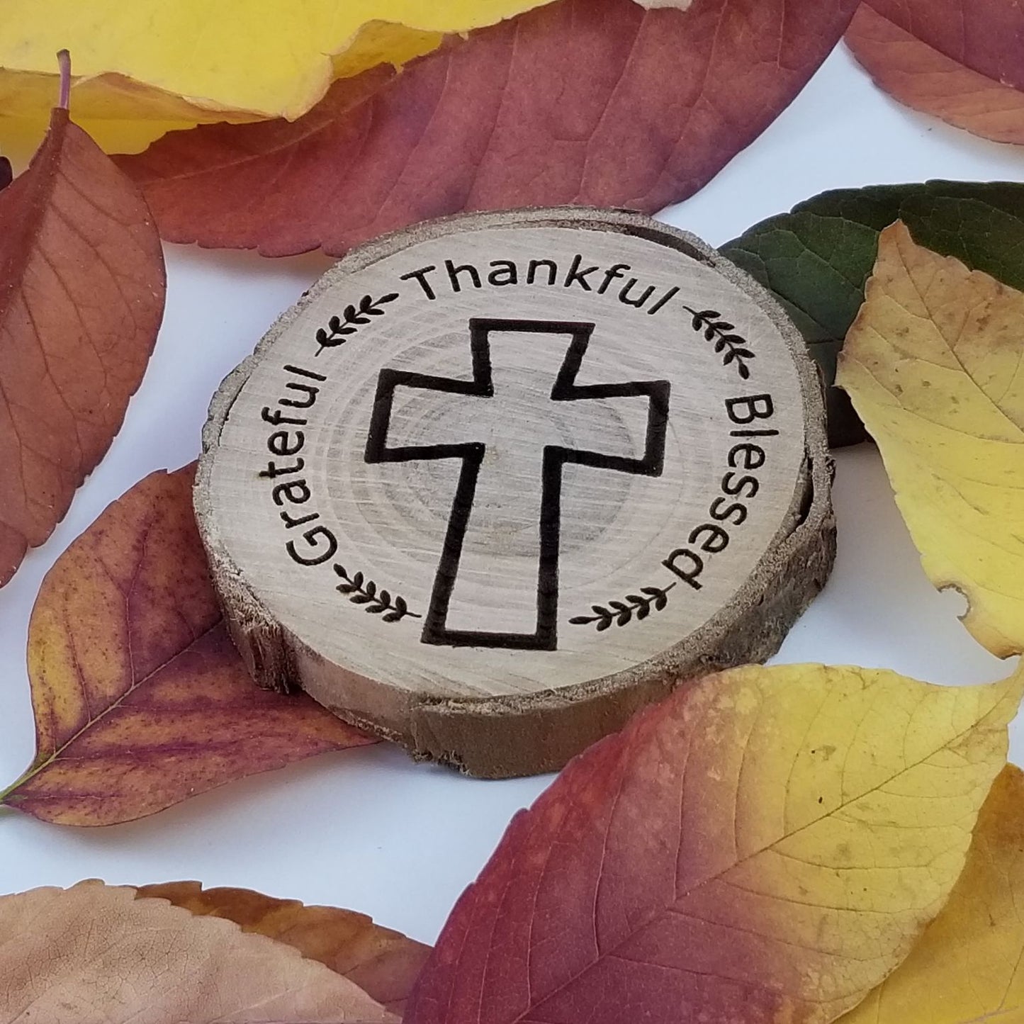 Wooden round plaque with cross design and engraved text on a background of autumn leaves.