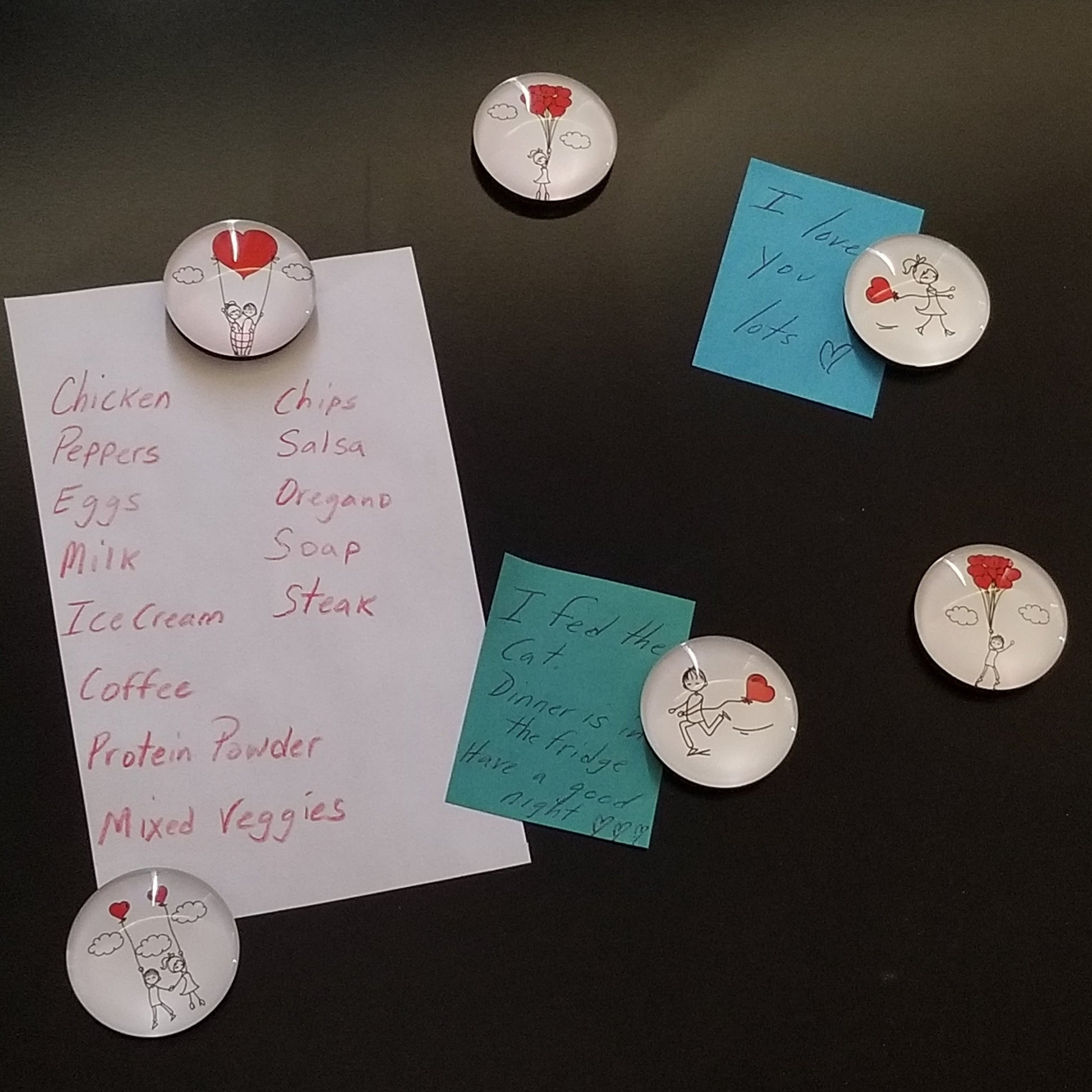 A set of round magnets with a 'Love' theme, featuring cartoonish designs of hearts and a smiley face, placed on a surface with a list of food items written on it.