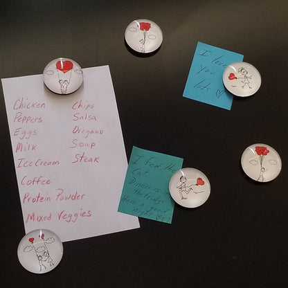 A set of round magnets with a 'Love' theme, featuring cartoonish designs of hearts and a smiley face, placed on a surface with a list of food items written on it.
