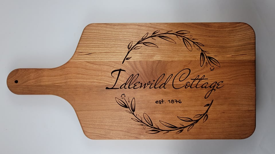 A wooden cutting board with a personalized engraving that includes a laurel wreath and the text 'Idlewild Cottage'.