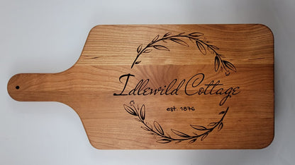 A wooden cutting board with a personalized engraving that includes a laurel wreath and the text 'Idlewild Cottage'.