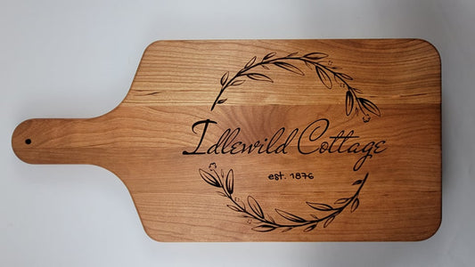 A wooden cutting board with a personalized engraving that includes a laurel wreath and the text 'Idlewild Cottage'.